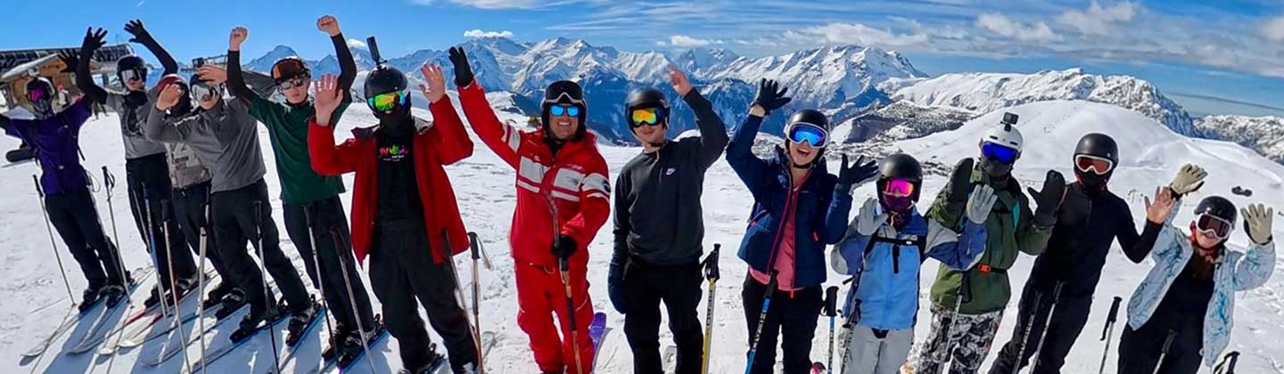 A group of student skiers on the top of a mountain with their ski instructor, smiling and with their hands in the air.